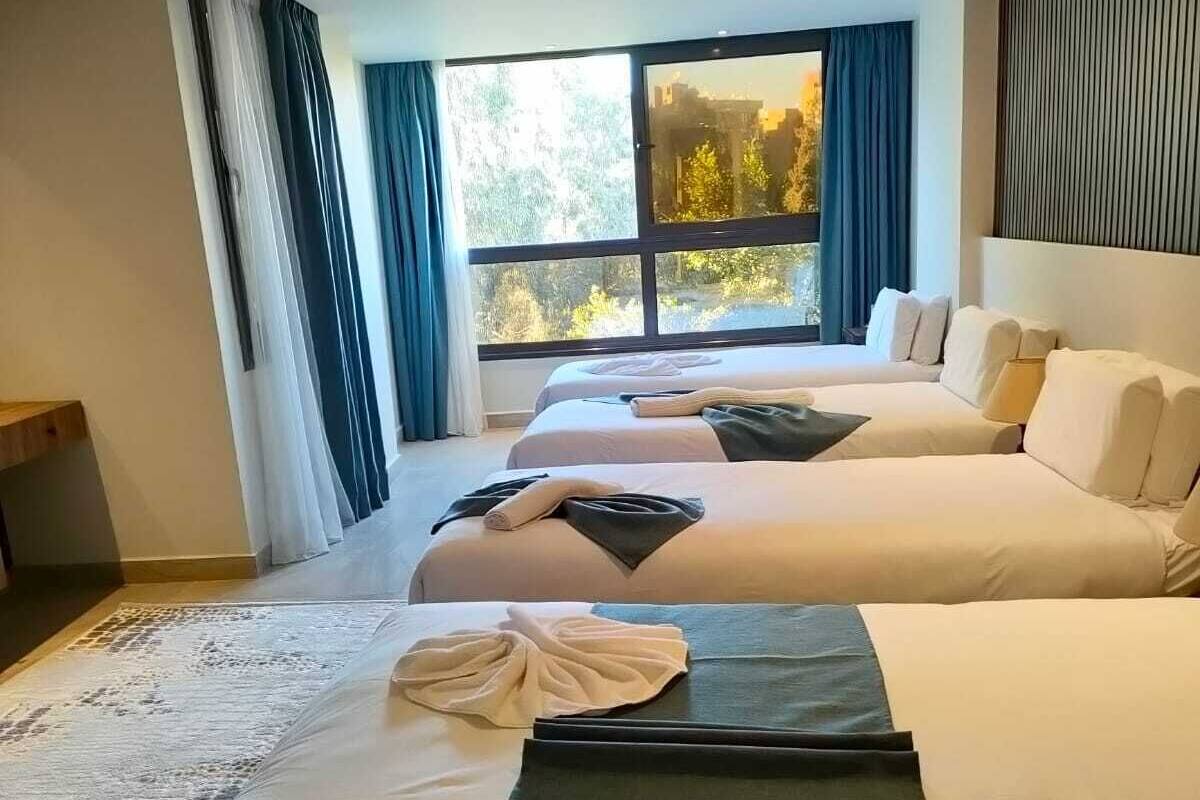 Quarto de hotel moderno com duas camas, uma janela ampla com vista para árvores e cabeceira de madeira.