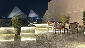 Un área con mesas y sillas al aire libre, un edificio moderno con un techo de forma piramidal, y un cielo nocturno.