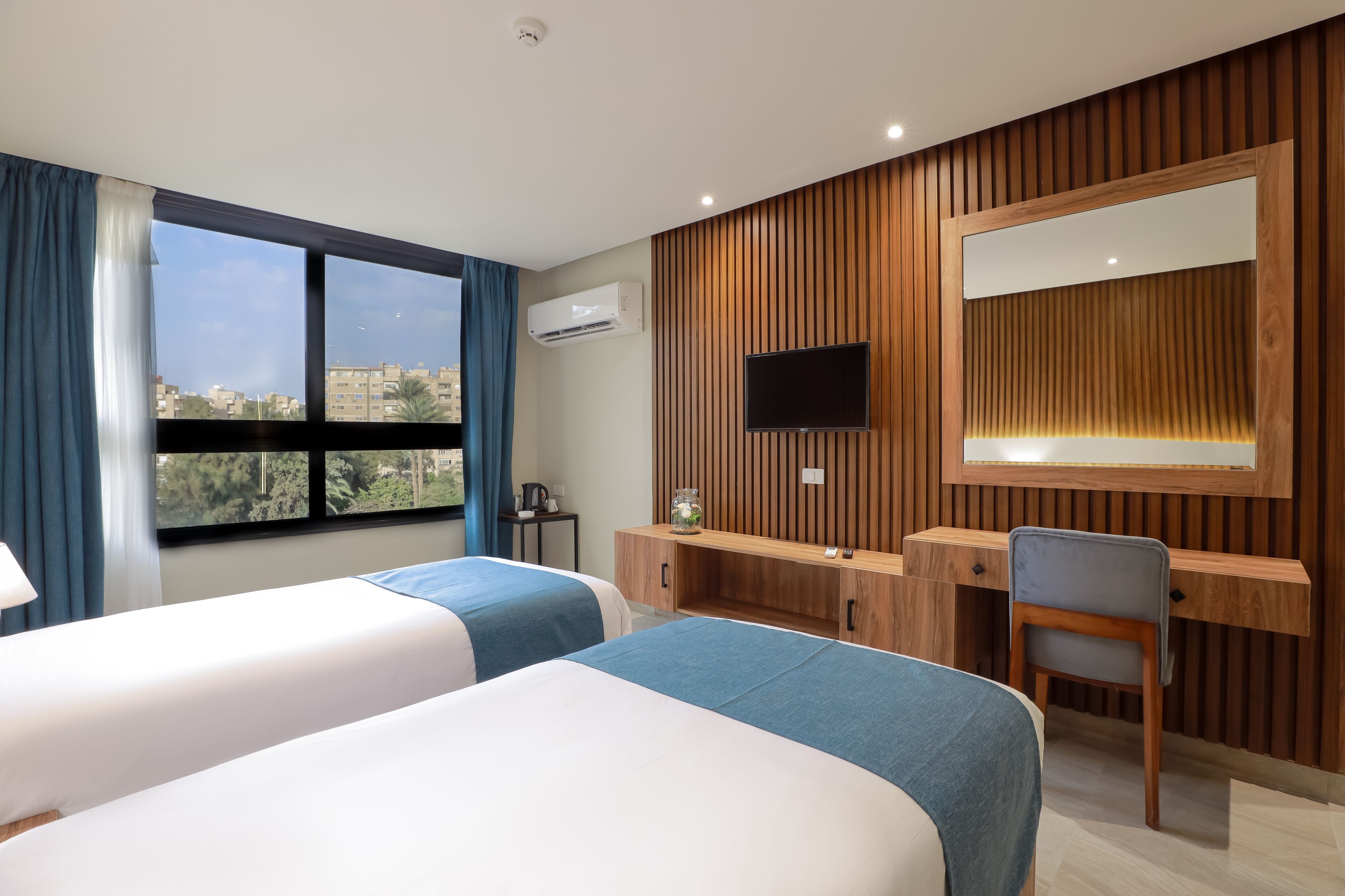 Een hotelkamer met twee bedden, een bureau, een stoel, een televisie en een groot raam met uitzicht op gebouwen en bomen.