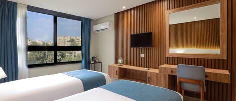 Een hotelkamer met twee bedden, een bureau, een stoel, een televisie en een groot raam met uitzicht op gebouwen en bomen.