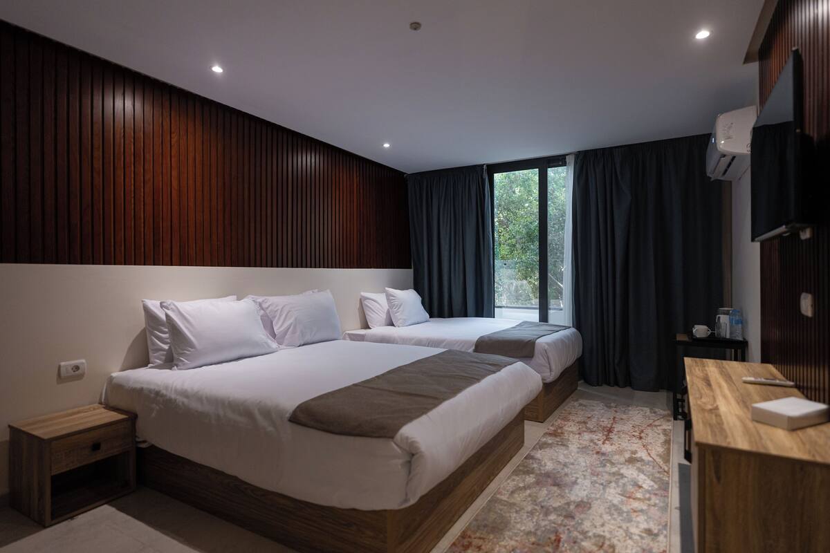 Quarto de hotel moderno com duas camas, painel de cabeceira de madeira, escrivaninha e uma grande janela com cortinas.