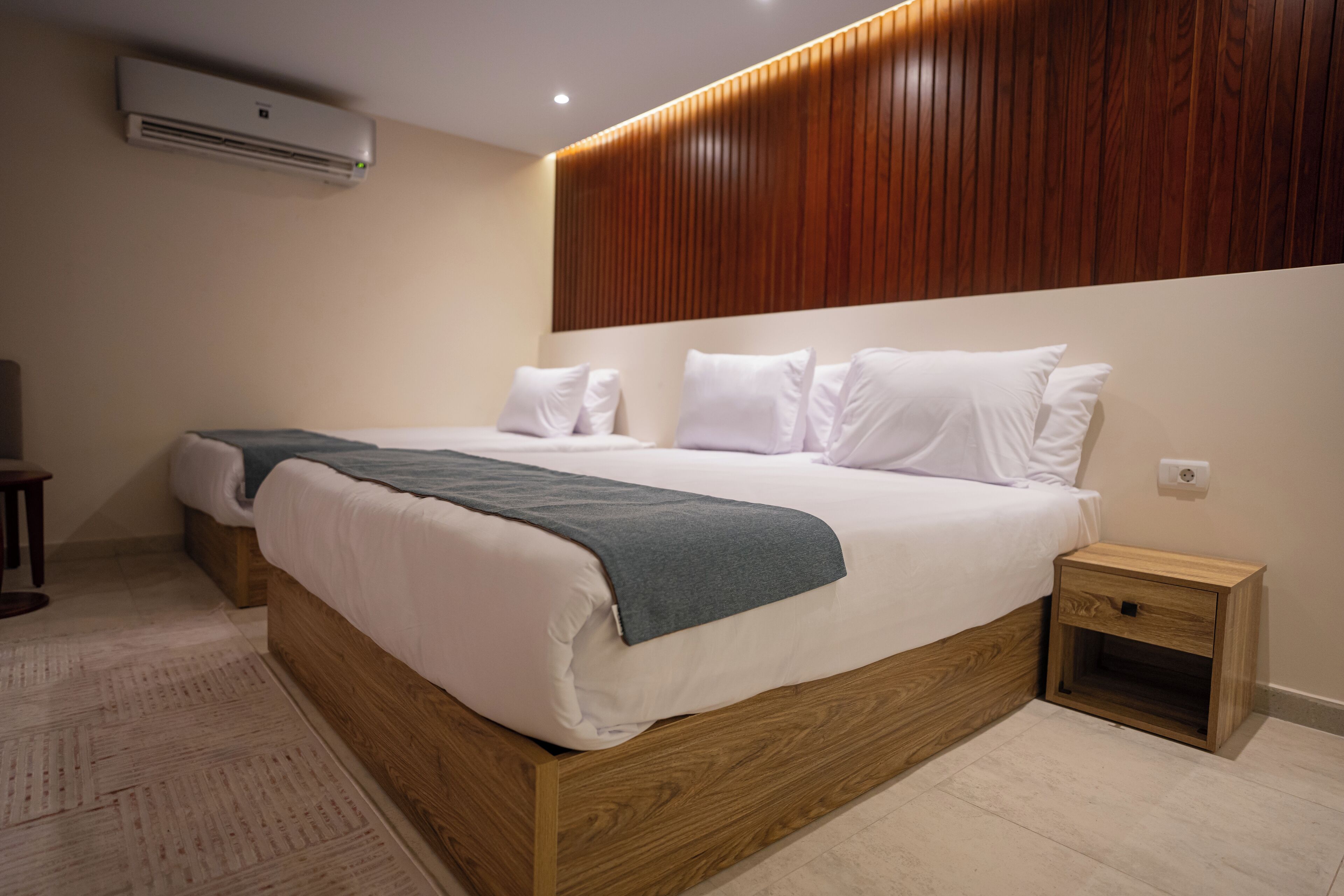 A bedroom with a wooden bed, white bedding, a bedside table, and an air conditioning unit.