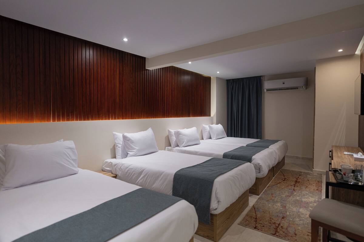 Quarto de hotel com três camas, cabeceira de madeira, unidade de ar condicionado, escrivaninha com cadeira e tapete estampado.