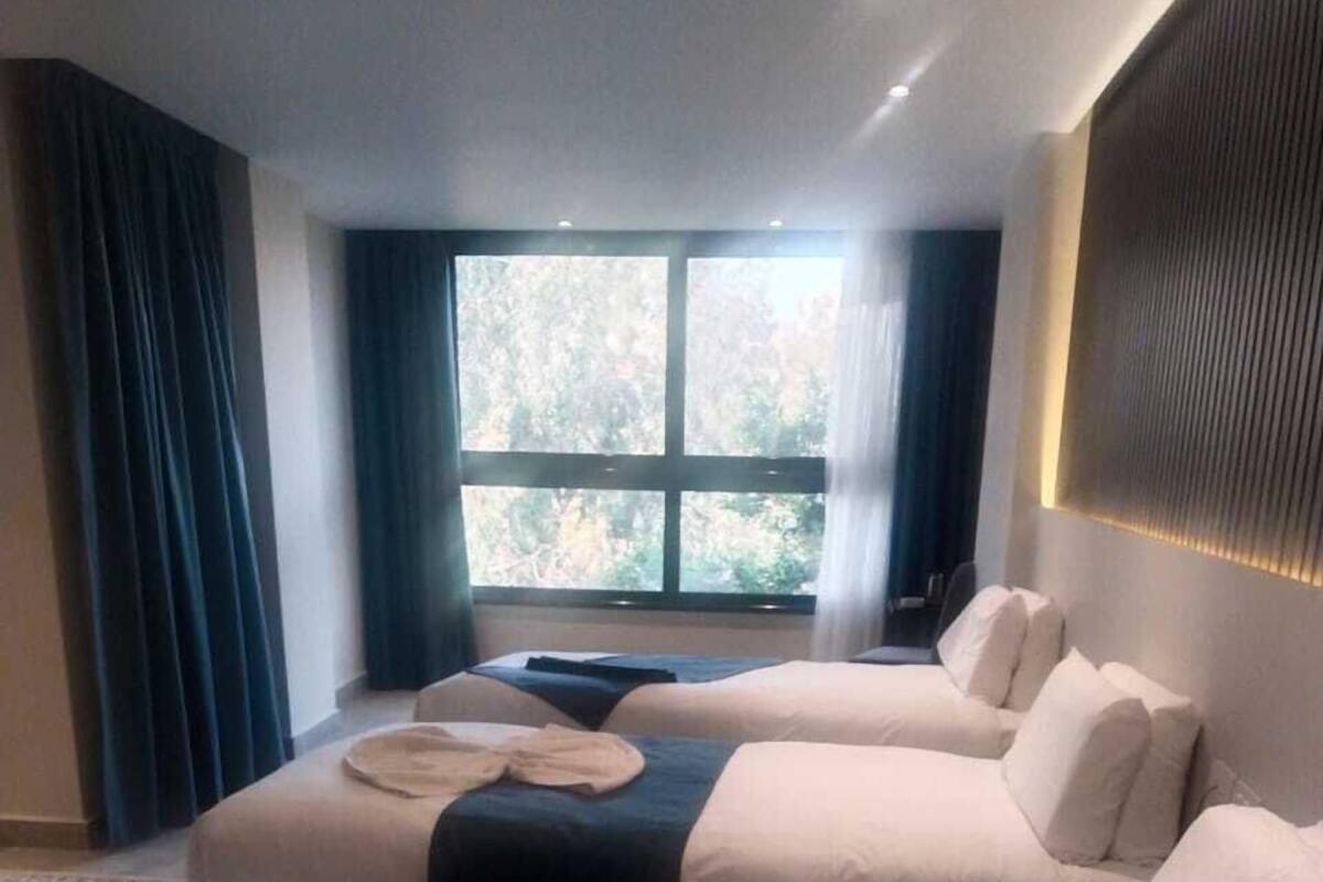 Quarto de hotel com duas camas, uma janela grande com cortinas e vista para árvores.