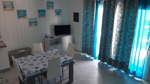 Una habitación con una mesa de comedor, sillas, un televisor sobre un mueble bajo y una ventana con cortinas florales.