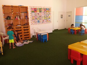 Un aula con una estantería de madera, un muro decorado con dibujos infantiles y suelo cubierto de alfombra.