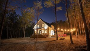 Ett modernt hus med en stor veranda på framsidan, omgivet av träd och upplyst av utomhusbelysning.