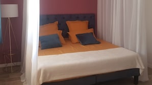 Ein Bett mit dunklem Kopfteil, orange- und blaue Kissen und einer weißen Tagesdecke.