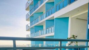 Ein Balkon mit Holztisch und -stühlen, einem Blumentopf und Blick auf ein blaues Gebäude sowie Grünflächen.