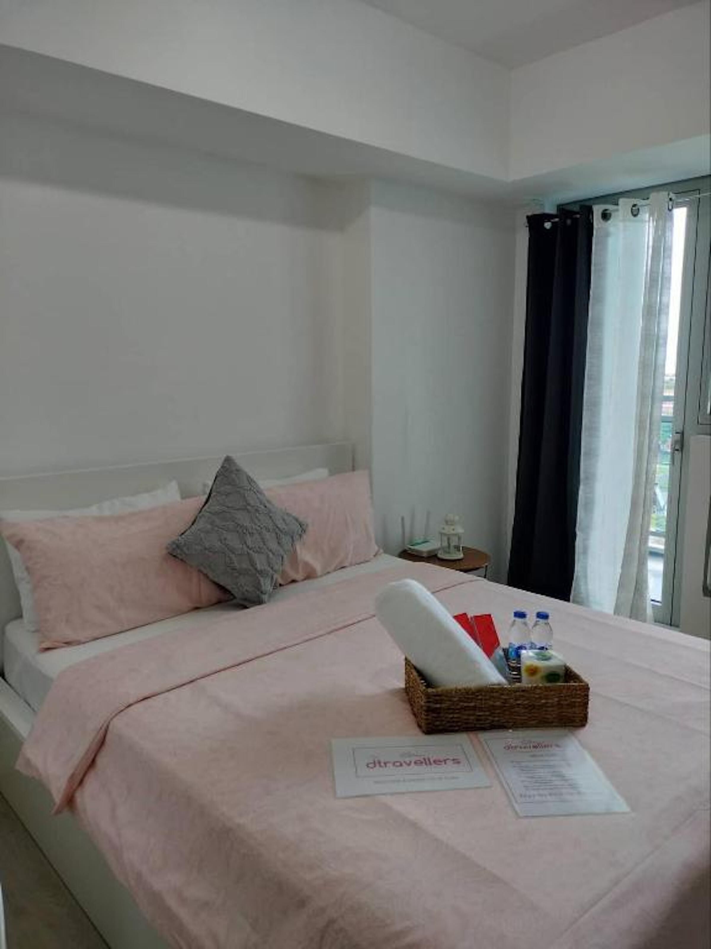 Una cama bien hecha con un edredón y almohadas rosas, una cesta de mimbre con artículos de aseo y un cartel que dice 'dtravellers' sobre la cama.