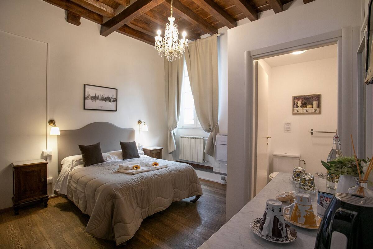 A bedroom with a bed, bedside lamps, a chandelier, a radiator, and a view of the bathroom.