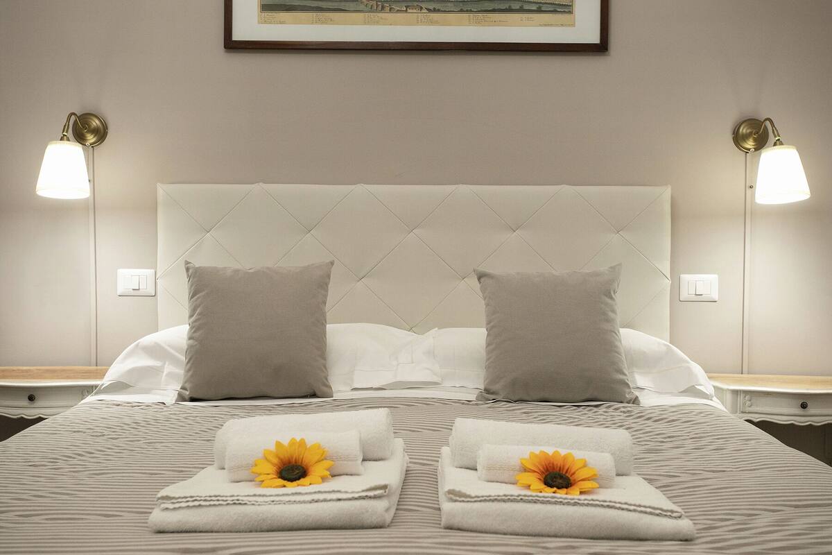 A neatly made bed with a tufted headboard, two pillows, and two folded towels with sunflowers on a striped bedspread.