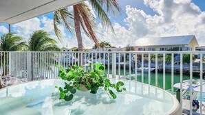 Een balkon met een glazen tafel, een bloempot en uitzicht op een jachthaven met boten en een huis op de achtergrond.