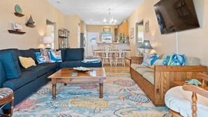 A living room with a sofa, coffee table, and patterned rug. There is a dining area with chairs and a table. - Ginger Mermaid / 916 E Dolphin Drive (Oak Island)