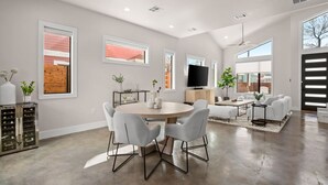 A spacious living room with a large round table, white chairs, a flat-screen TV, and a large window. - Grand Oasis At E.Austin W/ Pool (Austin)