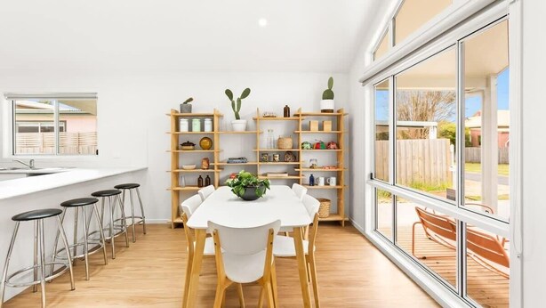 Una cocina moderna con encimera blanca, taburetes de madera y una mesa de comedor con sillas. Hay estanterías empotradas de madera con varios objetos y un gran ventanal con vistas al exterior.
