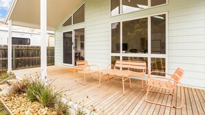 Un patio techado al aire libre con piso de madera, una mesa y sillas de metal, y un amplio ventanal corredizo.