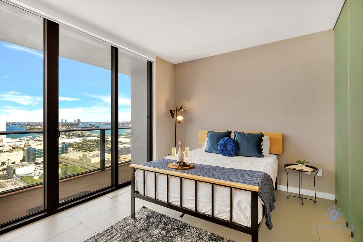 Un dormitorio moderno con un ventanal grande, una cama con cabecera de madera y una mesita de noche pequeña.