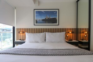 Habitación de hotel con una cama grande, dos mesitas de noche con lámparas, un cuadro en la pared y vistas de un paisaje urbano a través de la ventana.