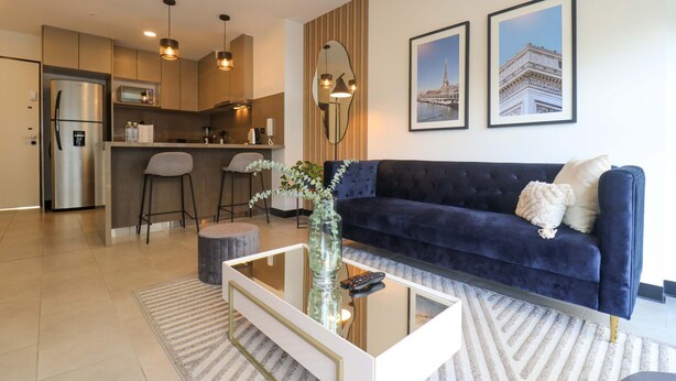A modern living room with a dark blue sofa, a glass coffee table, and a kitchen in the background.