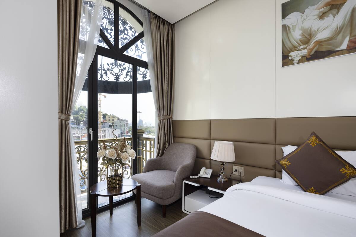 Una habitación de hotel con una cama, una silla, una mesita con un jarrón de flores, una mesita de noche con una lámpara y un cuadro en la pared.