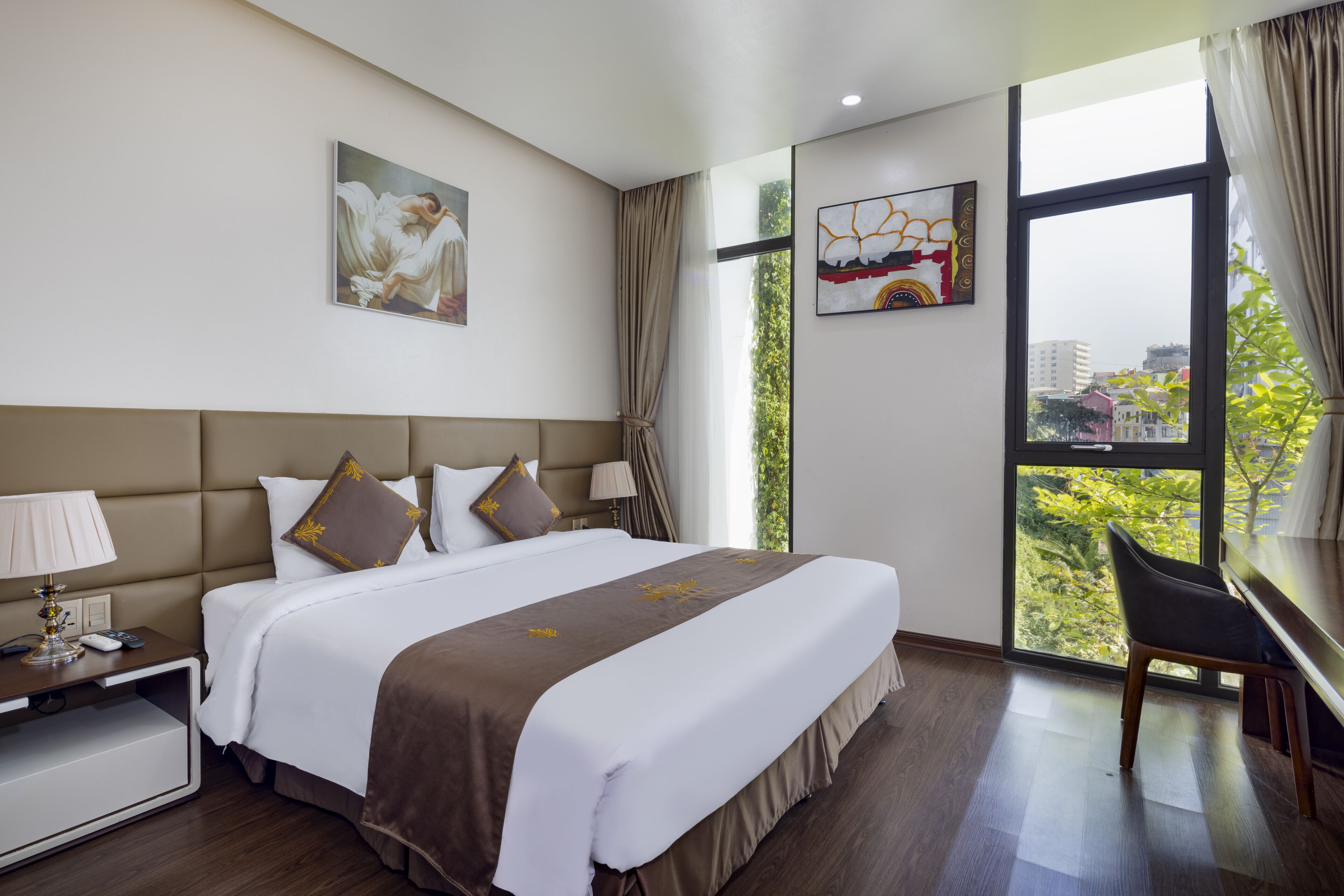 A hotel room with a large bed, a desk with a lamp, and a view of greenery through the window.