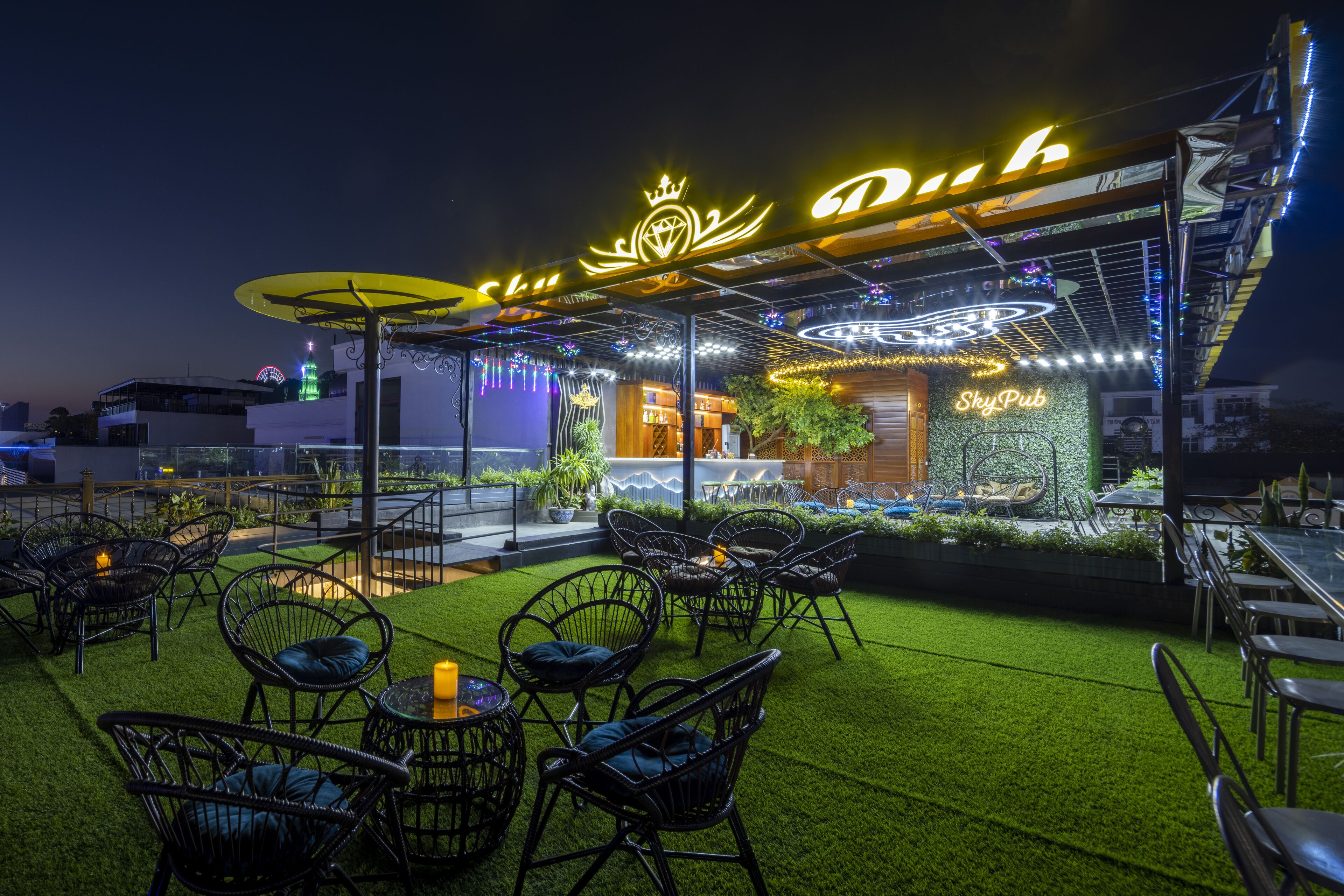 An outdoor seating area with artificial grass and wicker chairs, a neon sign reading "SkyPub," and a modern building facade in the background.