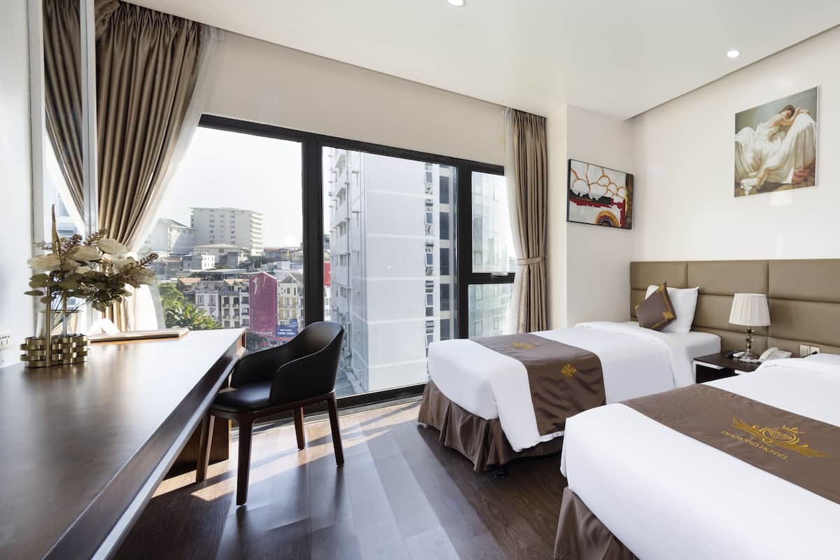 Habitación de hotel con dos camas, un escritorio y una silla. Cuenta con un ventanal con cortinas, un cuadro en la pared y vistas a edificios.