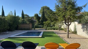 A swimming pool with a surrounding green lawn, two black and two yellow chairs, and a wooden shed in a garden setting.