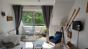 A modern living room with a sofa, armchair, and a small table. There is a balcony with a view of greenery.