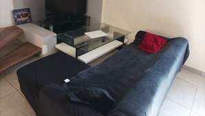 Ett vardagsrum med ett soffbord i glas, en svart soffa med en röd kudde, en tv och en inramad tavla på väggen.