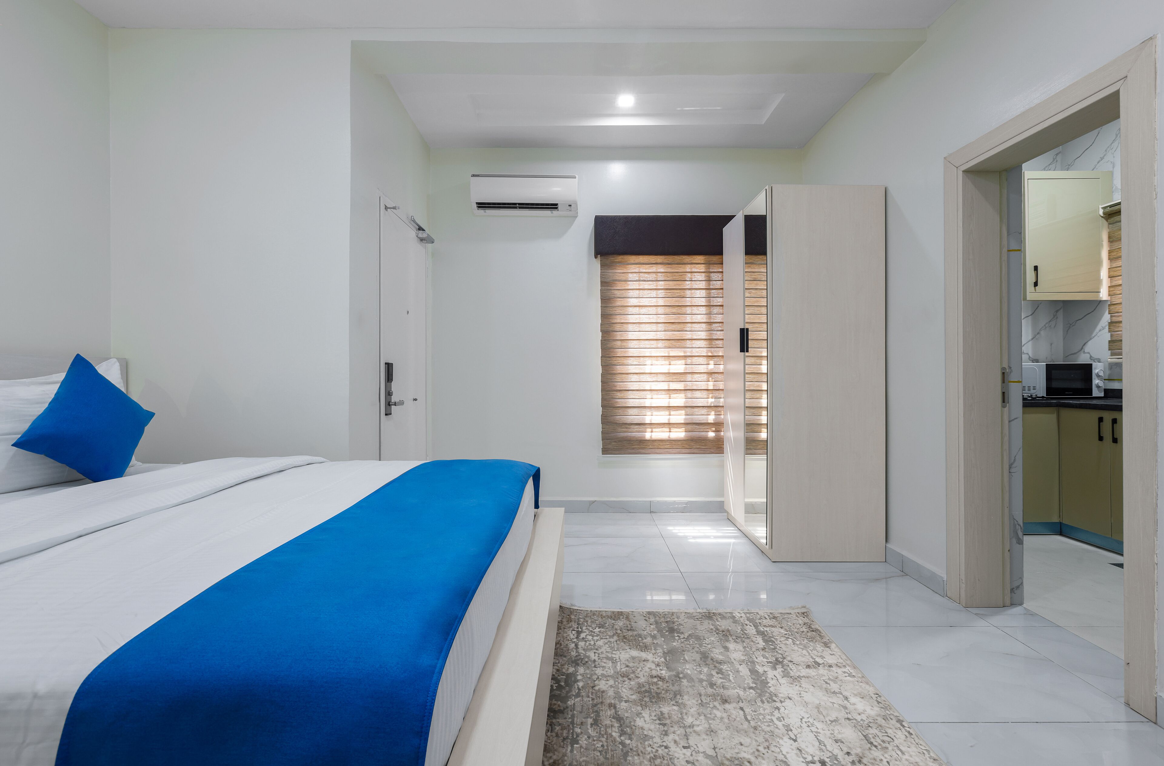 A modern bedroom with a bed, a blue accent pillow, a white air conditioner, and a view into the kitchen area.