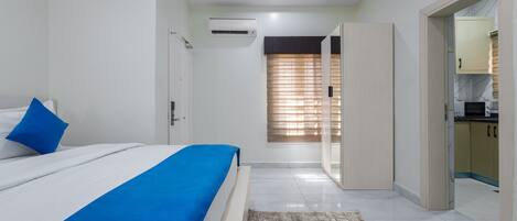 A modern bedroom with a bed, a blue accent pillow, a white air conditioner, and a view into the kitchen area.