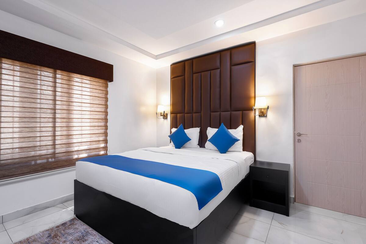 A modern hotel room with a large bed, a wooden headboard, bedside lamps, and a door.