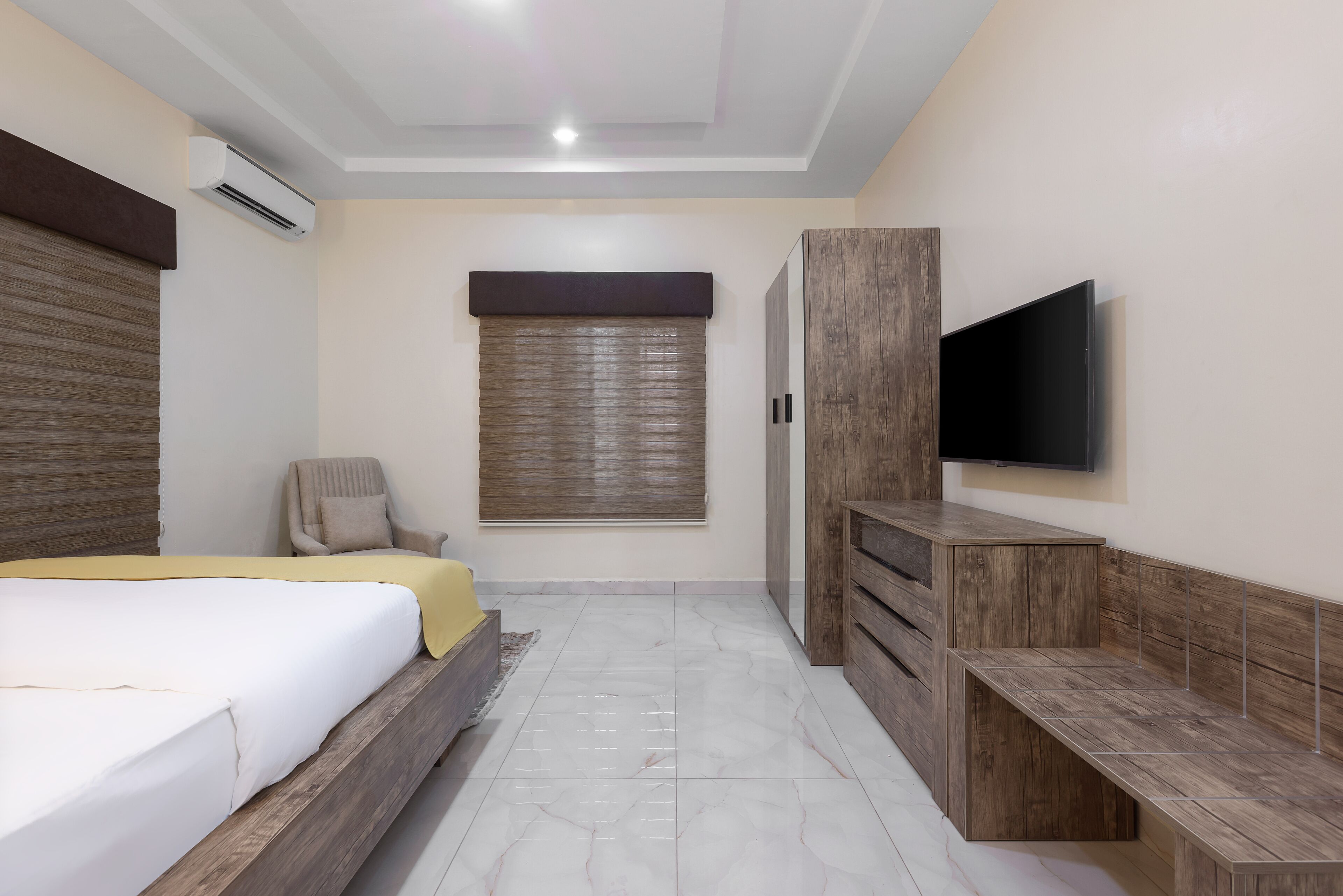 A bedroom with a bed, a TV mounted on the wall, a wooden wardrobe, and a window with blinds.