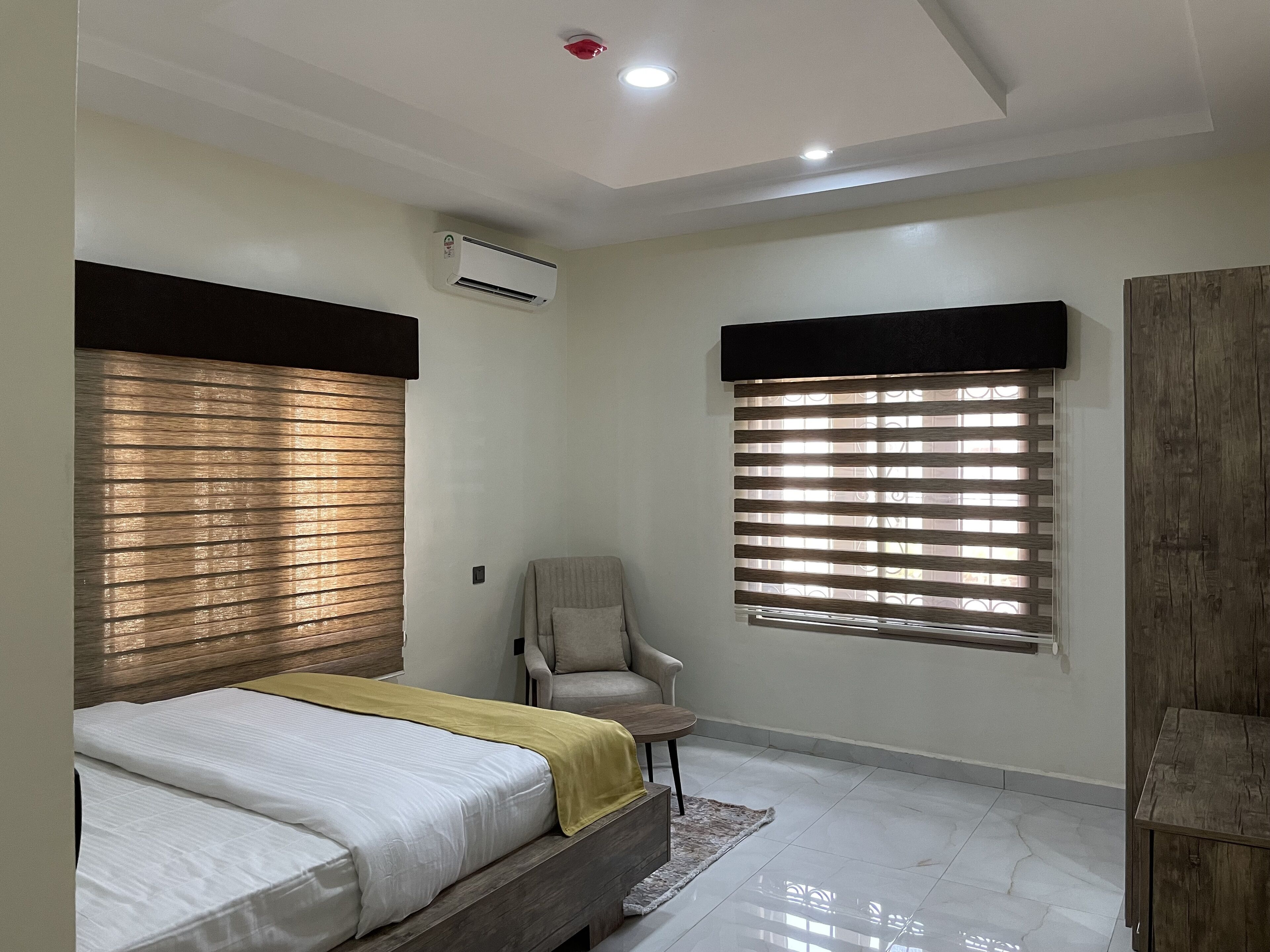 Un dormitorio con cama, una silla, ventana con persianas, unidad de aire acondicionado y una lámpara en el techo.