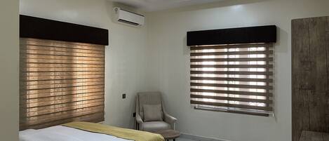 Une chambre à coucher comprenant un lit, une chaise, une fenêtre avec des stores, un appareil de climatisation et un luminaire au plafond.