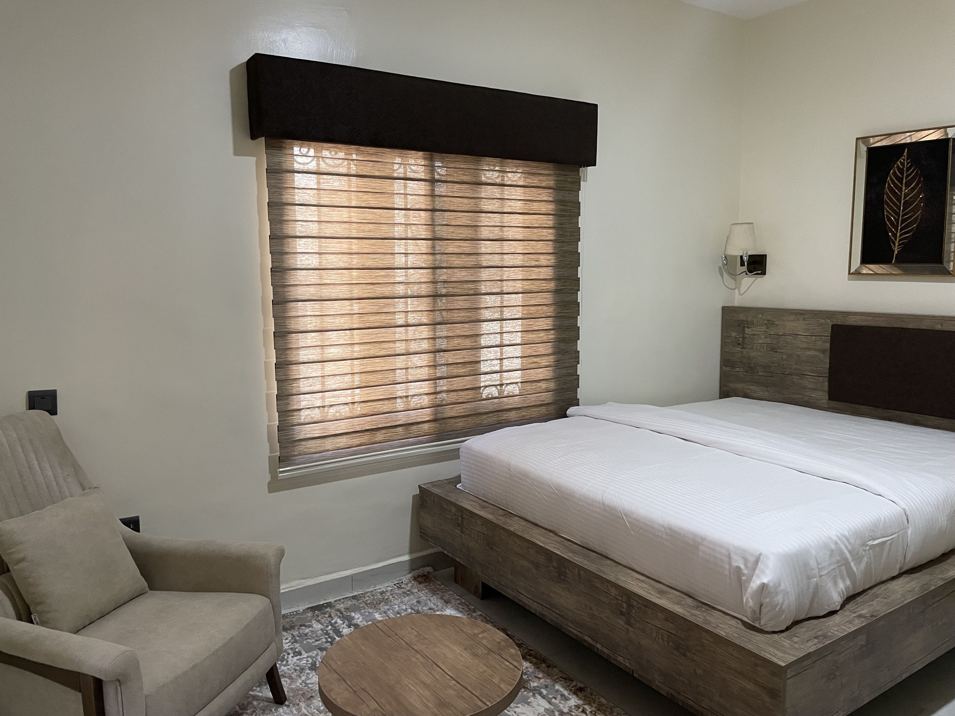 Een slaapkamer met een bed, een stoel, een ronde houten tafel en een raam met jaloezieën.