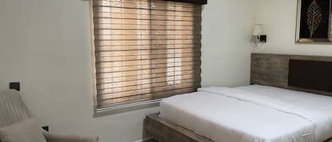 A bedroom with a bed, a chair, a round wooden table, and a window with blinds.