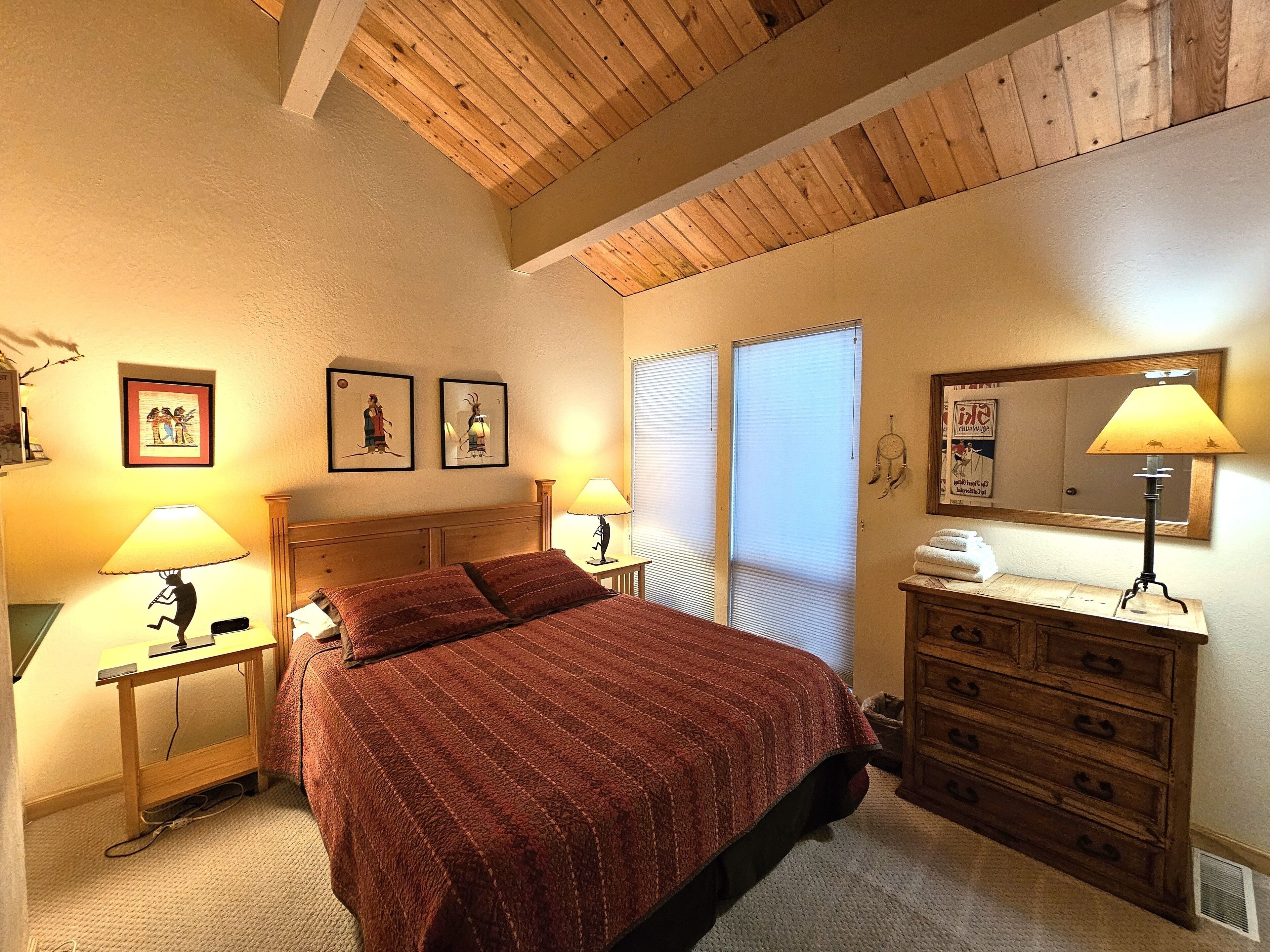 Un dormitorio con techo de madera, una cama con manta roja, dos lámparas de noche, un aparador y una pared con cuadros enmarcados.