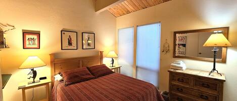 Un dormitorio con techo de madera, una cama con manta roja, dos lámparas de noche, un aparador y una pared con cuadros enmarcados.