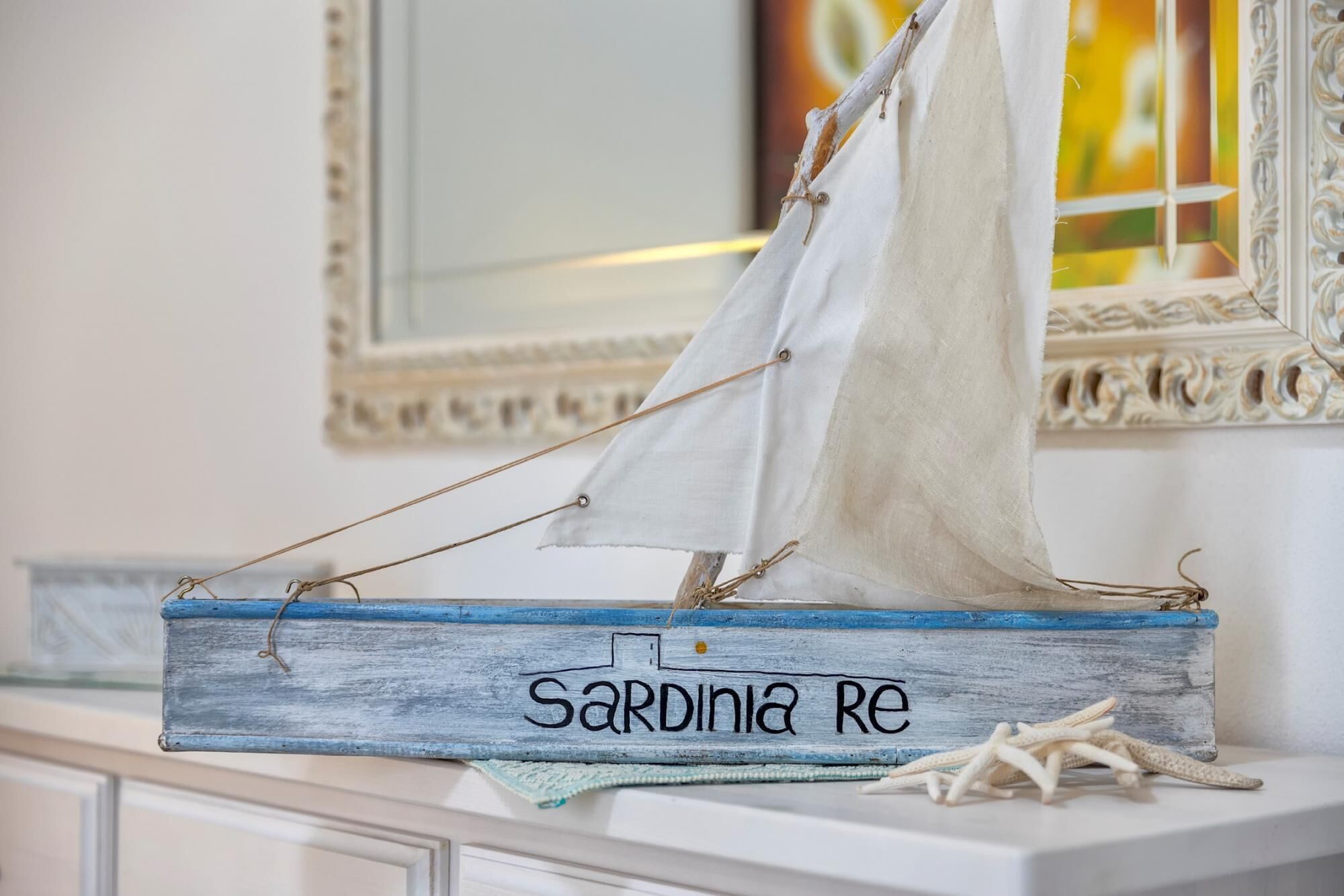 A model sailboat named 'Sardinia Re' on a wooden surface with a mirror and decorative elements in the background.