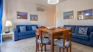 A living room with a blue sofa, a wooden dining table, and a white pendant light. There are three framed pictures on the wall. - Casa Grace in Alghero (Alghero)