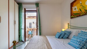 A bedroom with a bed, pillows, a painting, and a view of buildings through the window. - Casa Grace in Alghero (Alghero)