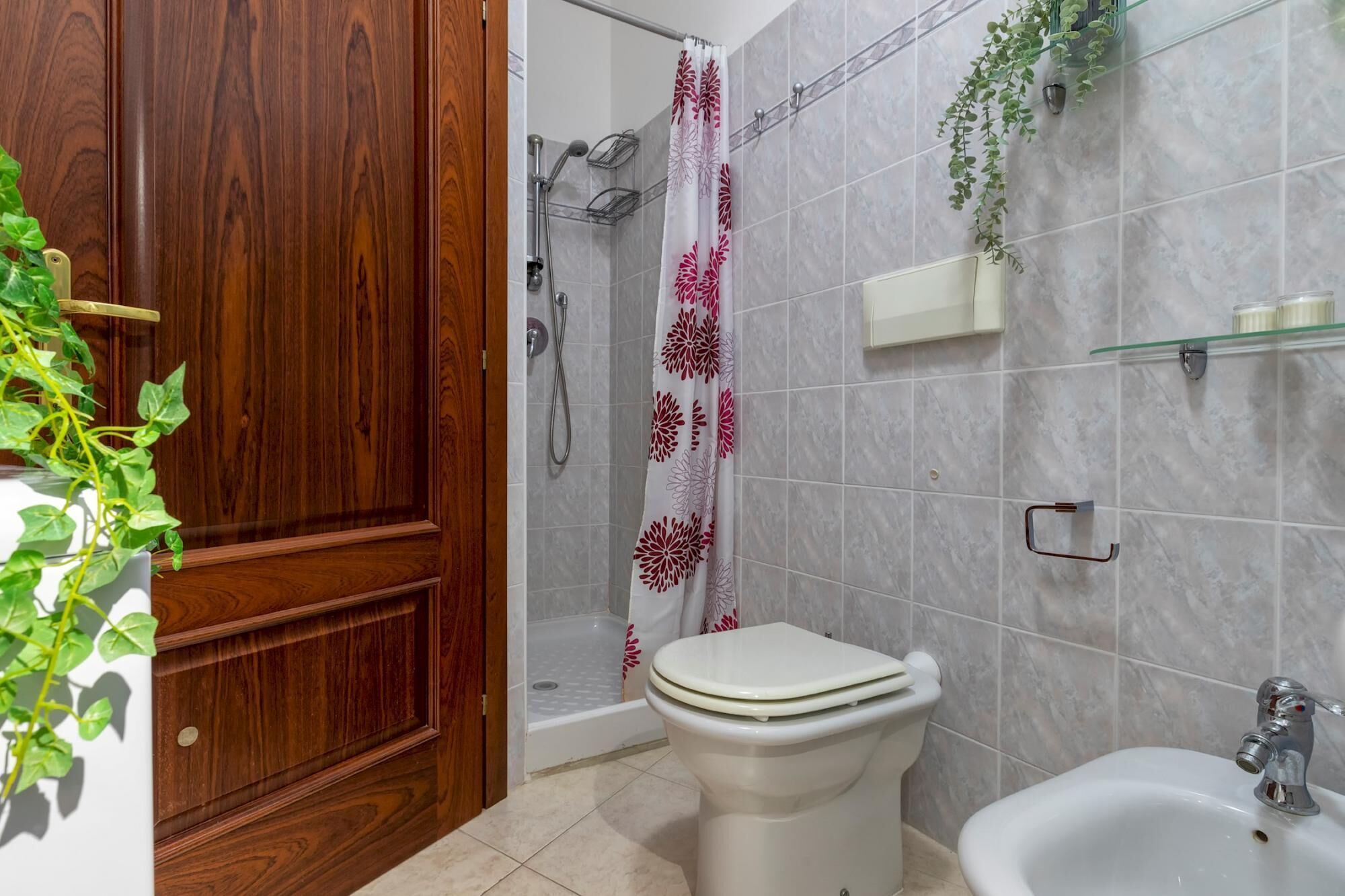 A bathroom with a wooden door, a shower with a floral curtain, a toilet, a bidet, and a shelf with toiletries.