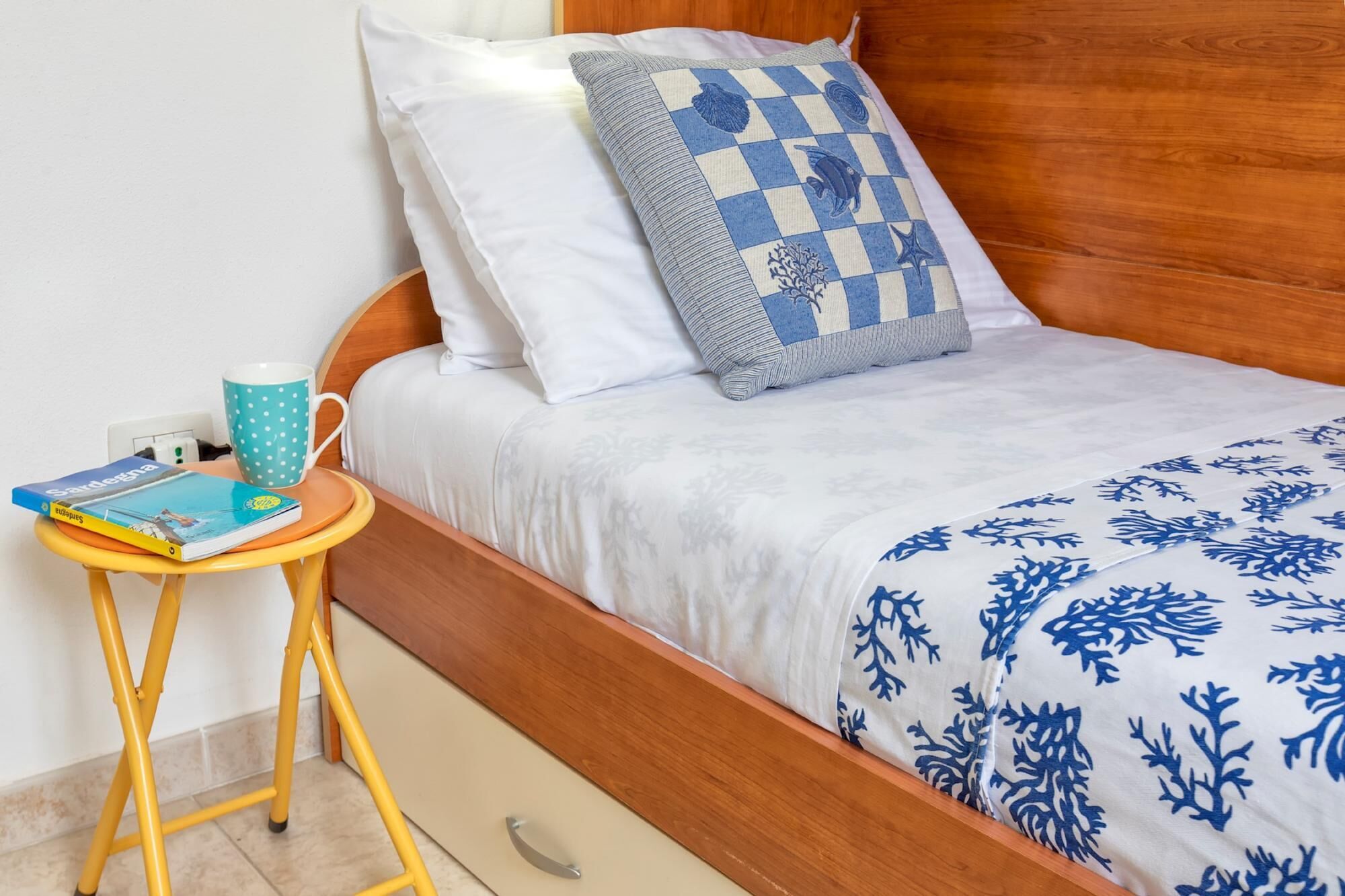 Een net opgemaakt bed met een blauw-witte sprei, een houten hoofdbord en een klein nachtkastje met een puntjesmok en een boek.