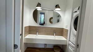 Un baño pequeño con un lavabo blanco, un espejo, dos luces empotradas en la pared, una lavadora y un estante de madera.