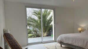 Un dormitorio con cama, mesita de noche, lámpara, silla de madera y puerta corrediza de vidrio que da a un jardín con palmeras.