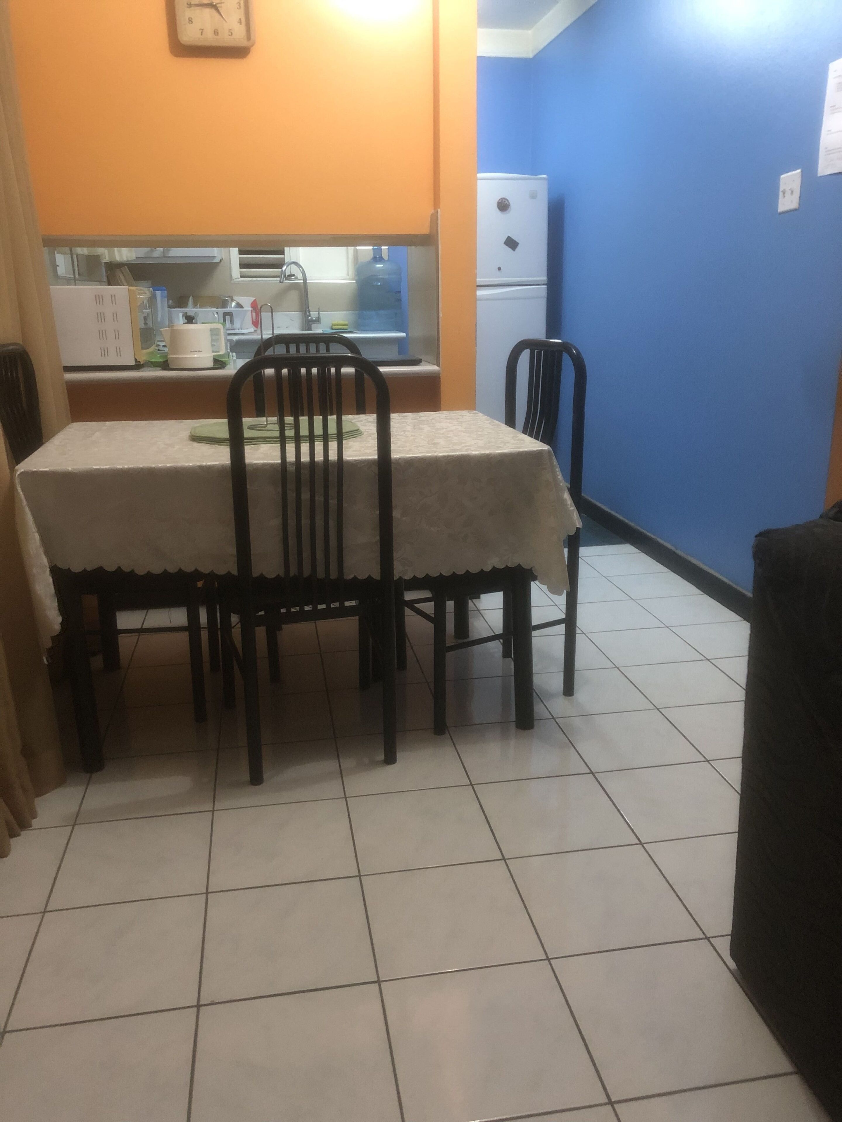 A dining area with a table and chairs, a kitchen in the background, and a blue wall.