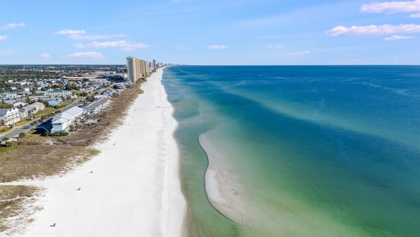 Ein Luftbild einer Küstenregion mit einem breiten Sandstrand, klarem, blauem Wasser und einer Reihe von Gebäuden entlang des Ufers.